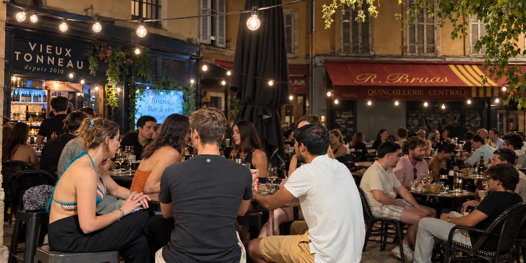 Une tablée d’amis sur des tabourets, en terrasse le soir, sous les guirlandes et l’enseigne du Vieux Tonneau.