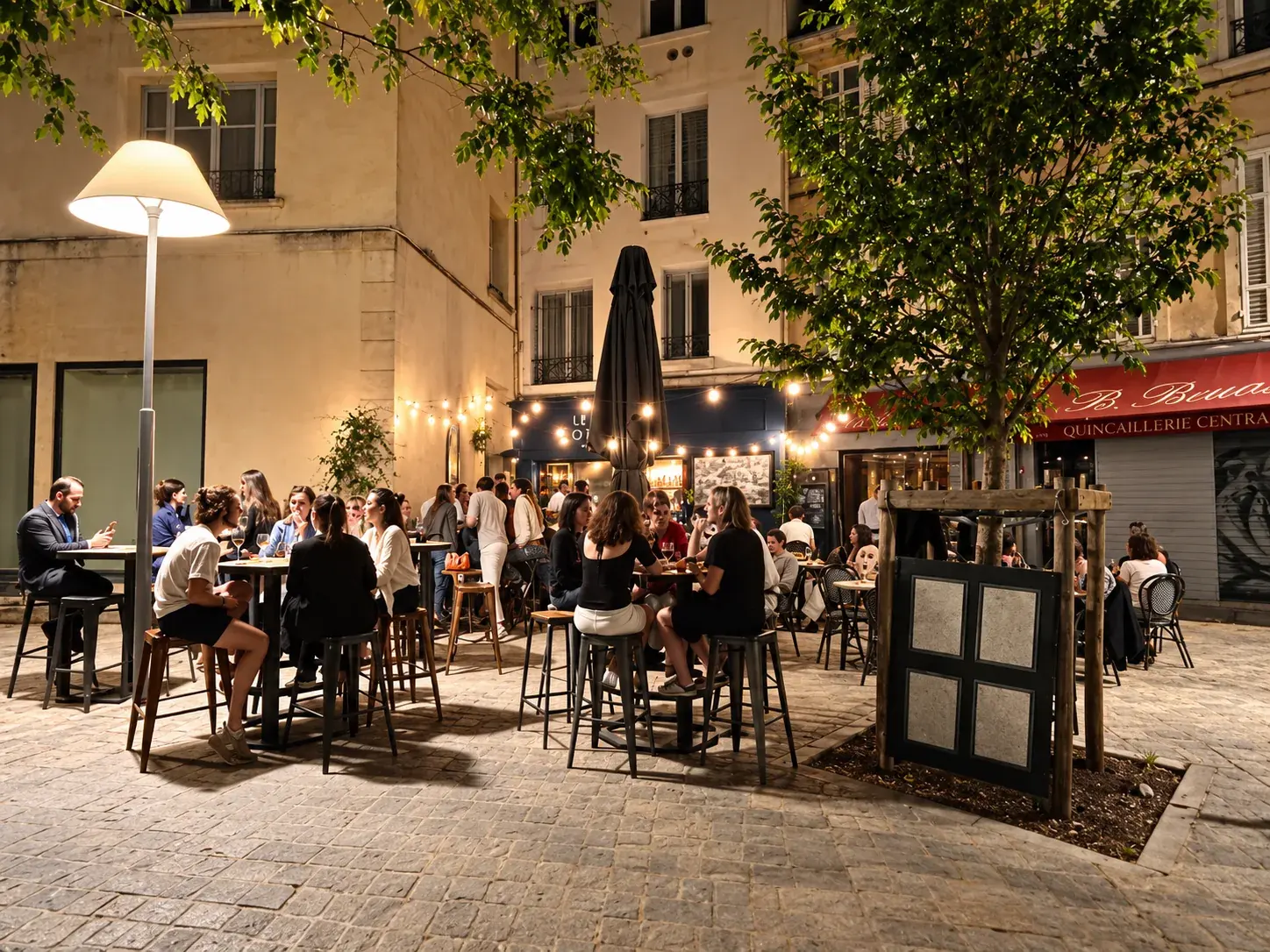 La placette de nuit : les tables de la terrasse du Vieux Tonneau sous les guirlandes, avec l’arbre et le panneau de la carte.